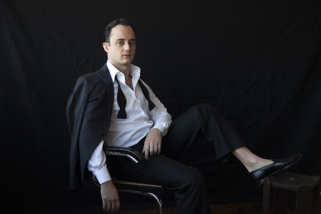 Portrait of Tolga seated in a dark suit against black, by Mathias Kessler